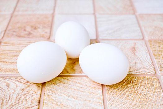 an oval or round object laid by a female bird, reptile, fish, or invertebrate, usually containing a developing embryo. Three chicken bird eggs on a wooden pink orange checkered surface.