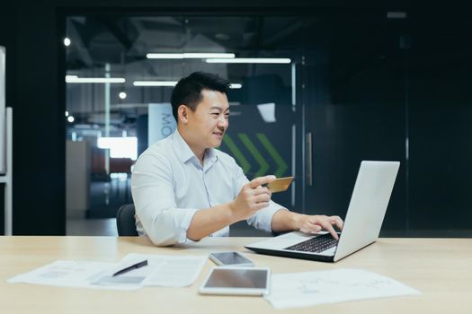 Cheerful and smiling asian businessman working in modern office, dark colors, making online purchase, using bank credit card and laptop, for online shopping