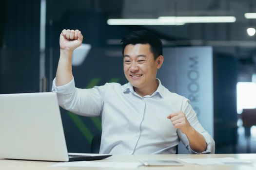 Cheerful and successful asian businessman dancing in office celebrating triumph and victory sitting at desk working with laptop