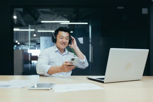 Businessman in office resting during work break, listening to music in big black headphones, Asian man using music app on phone