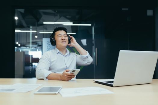 Businessman in office resting during work break, listening to music in big black headphones, Asian man using music app on phone