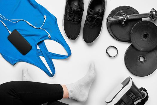 Flat lay gym equipment such as dumbbells, bottle of waters, smartphone with headphone, fitness bracelet and human legs in socks on white background. Fitness concept