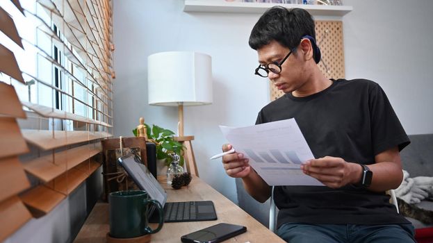 Young asian man holding financial graphs and statistics documents and working with computer tablet at home.