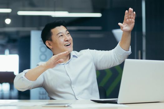 Cheerful and successful asian businessman dancing in office celebrating triumph and victory sitting at desk working with laptop