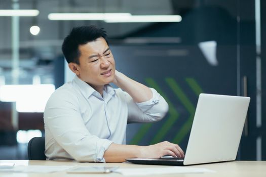Overtired asian worker in office, having severe neck pain, massaging with hand, businessman working in office with laptop