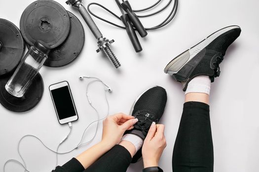 Young woman in sporting leggings laces sneakers, preparing for training. Accessories for sports, bottle of water, smartphone, headphones, fitness bracelet, dumbbells on white background flat lay top view. Fitness concept