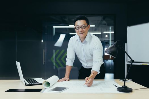 Portrait of successful and experienced Asian architect, man standing over table with plan drawing, smiling and looking at camera, working in modern design studio