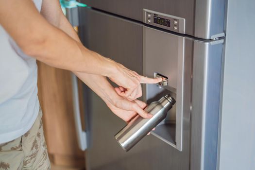 Male hand is pouring cold water and ice cubes in a metal bottle from dispenser of home fridge.