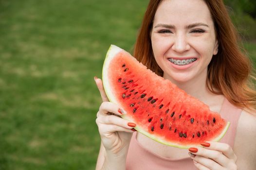 Beautiful red-haired woman smiling with braces and about to eat a slice of watermelon outdoors in summer.