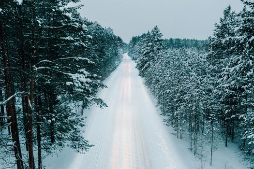Aerial drone view of snowy winter road in forest with light lines from car. Night time aerial view of snowy road in pine tree forest. Drone perspective view of snowy winter road surrounded fir forest