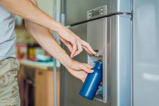 Male hand is pouring cold water and ice cubes in a metal bottle from dispenser of home fridge.
