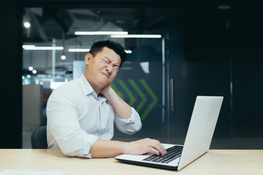 Overtired asian worker in office, having severe neck pain, massaging with hand, businessman working in office with laptop
