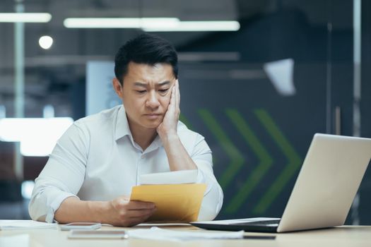 Sad over thinking asian businessman reading bad news letter from bank, male boss in shirt working in office