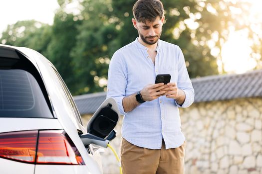 Caucasian bearded man using smart phone and waiting power supply connect to electric vehicles for charging the battery in car. Plug charging an Electric car from charging station.