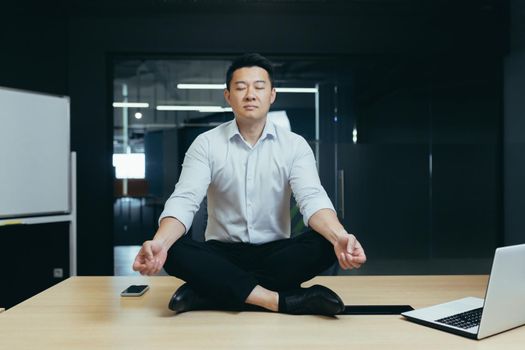 Asian boss businessman meditating in office, man sitting in lotus position on desk with closed eyes, thinking about important decision