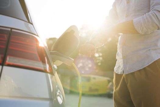 Unrecognizable man disconnects charging cable to to electric vehicle using smartwatch. Male hand unplugs power connector into EV car and charges batteries, uses smart watch to disconnect charging