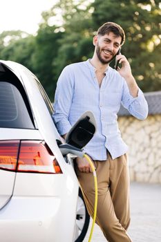 Portrait of handsome joyful 30-aged man with beard which has mobile conversation while luxurious car charging battery on specially equipped charging station.