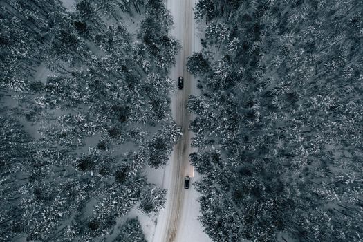 Night time aerial view of snowy road in pine tree forest in winter season. Drone top down view of snowy winter road surrounded pine and fir forest and two cars with light