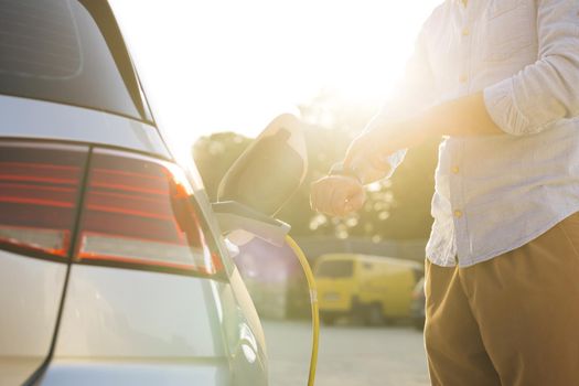 Businessman plugging in charging cable to to electric vehicle. Male hand inserts power connector into EV car and charges batteries, uses smartwatch for activates start charging.