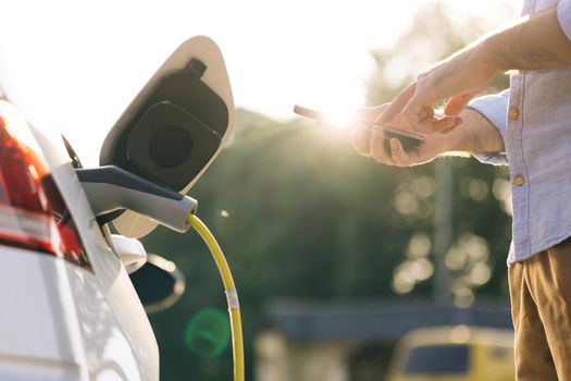 Male unplugging in power cord to electric car using app on smartphone. Businessman charging electric car at outdoor charging station Unrecognizable man unplugging electric car from charging station.