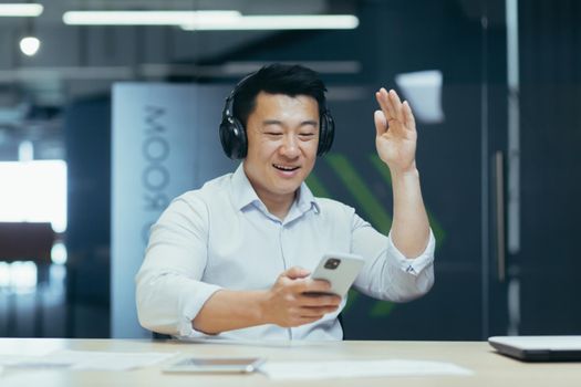 Businessman in office resting during work break, listening to music in big black headphones, Asian man using music app on phone