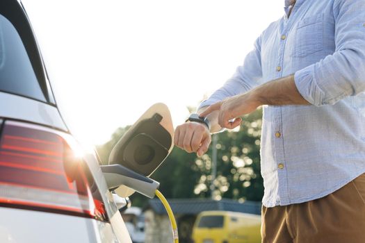 Male hand unplugs power connector into EV car and charges batteries, uses smart watch to disconnect charging. Man disconnects charging cable to to electric vehicle using smartwatch.