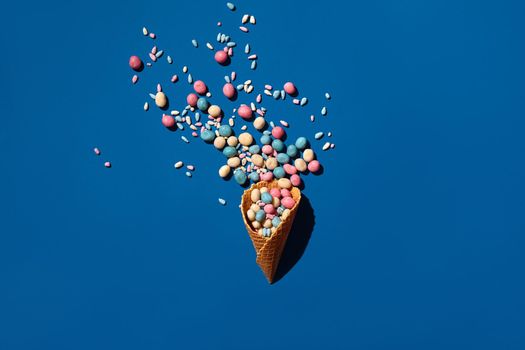 Flat lay photo of a heap of candy dragee that is sprinkled from a waffle cone on blue background from above