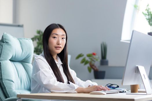 Close-up photo. Portrait of a young beautiful Asian woman secretary sitting at the desk at the computer in the office in business clothes, looking at the camera, smiling.