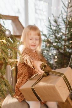 A little blonde girl is sitting on a wooden staircase in a Scandinavian interior decorated in a New Year's style. A child holds a Christmas gift in a craft package.