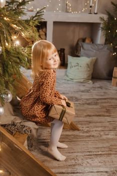 A little blonde girl is sitting on a wooden staircase in a Scandinavian interior decorated in a New Year's style. A child holds a Christmas gift in a craft package.