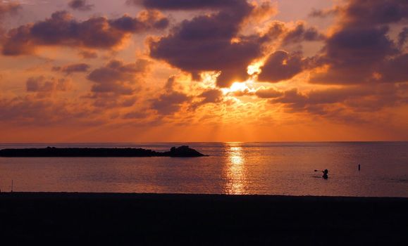 Sunset through the clouds at Pokai Bay light reflects on water and paddler in the water.