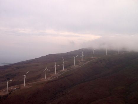 Aerial view of a line of Modern Windmills spin on hillside with windy road and cloudy skies on the island of Maui.