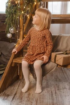 A little blonde girl is sitting on a wooden staircase in a Scandinavian interior decorated in a New Year's style. A child holds a Christmas gift in a craft package.