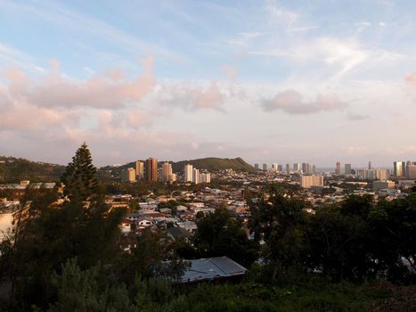 Punchbowl Crater, Nuuanu, and Honolulu Cityscape at dusk looking to the ocean from high up in the hills with cranes and modern skyscrapers mixed with houses, and other small buildings on a beautiful day. With trees in the foreground.