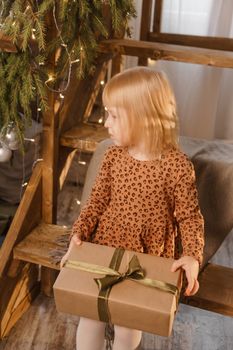 A little blonde girl is sitting on a wooden staircase in a Scandinavian interior decorated in a New Year's style. A child holds a Christmas gift in a craft package.