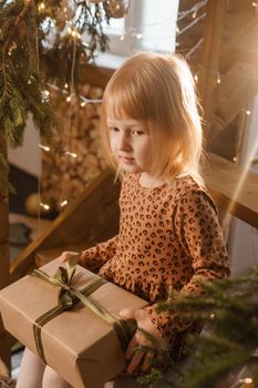 A little blonde girl is sitting on a wooden staircase in a Scandinavian interior decorated in a New Year's style. A child holds a Christmas gift in a craft package.