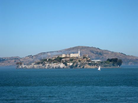 Fog rolls onto Alcatraz Island on a nice Day with a sail boat in front of it and Angel Island in the distance behind it. 