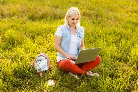 Portrait of pretty young woman sitting on green grass in park with legs crossed during summer day while using laptop for video call.