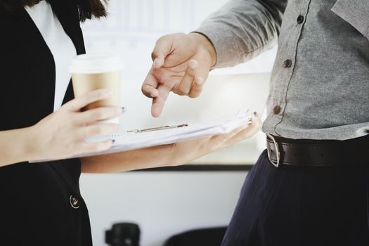 Focus on the man's hand pointing to the paperwork and chatting with a coworker who is having a cup of coffee and holding a budget document in the afternoon