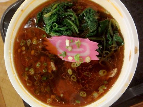 Oily Spicy Beef Ramen Soup in a bowl with onions, fish cake, and spinach on a plastic tray.