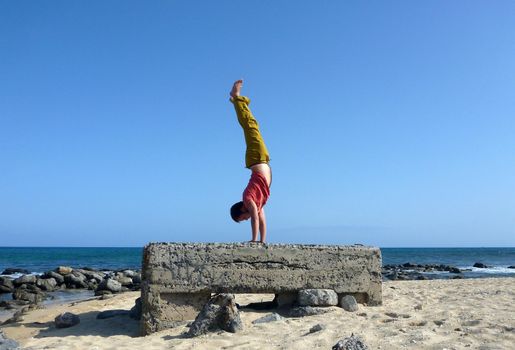 Man wearing a red t-shirt, long olive pants, and bare feet Handstands on top a old historic pillbox on the beach Sand Island Beach on Oahu, Hawaii