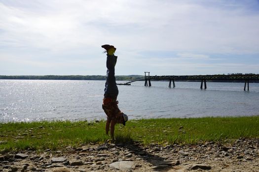 Man wearing a hat, aloha shirt, pants, and shoes Man Handstands on rocky grassy beach with Pier on Cousins Island, Maine.