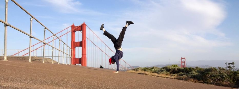 Man wearing hat, hoodie, long pants and shoes Handstands in front of the Golden Gate Bridge on the Marin side with San Francisco, California in the distance.