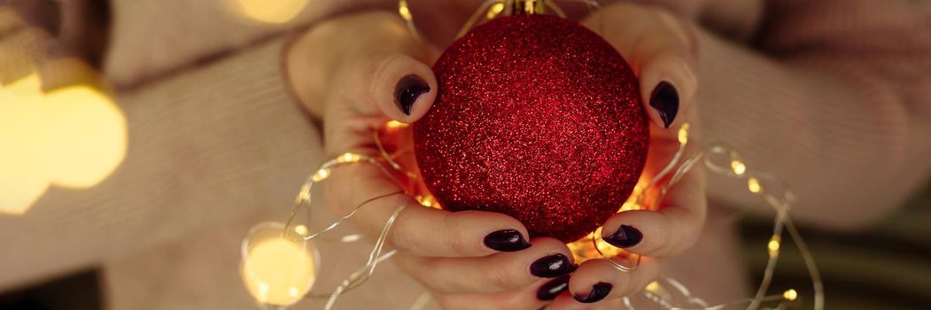 Women's hands in a sweater hold a Christmas ball with garland lights. New year and christmas concept. Copy space, web banner.