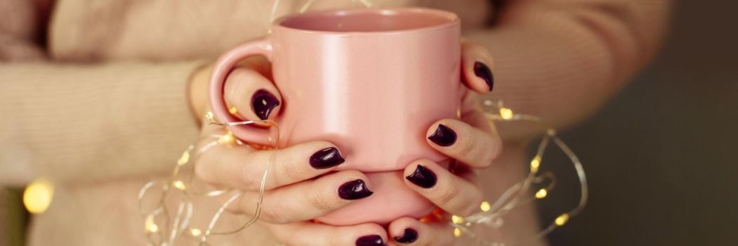 Female hands hold a cup of warm coffee, cocoa or tea with garland lights. Web banner, copy space.