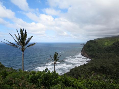 Pololu Valley cliffs and the dramatic northeastern coastline with small island outcroppings sitting in the waters offshore in the distance and the black sand beach at the mouth of the valley as wave break towards shore in Big Island, Hawaii.