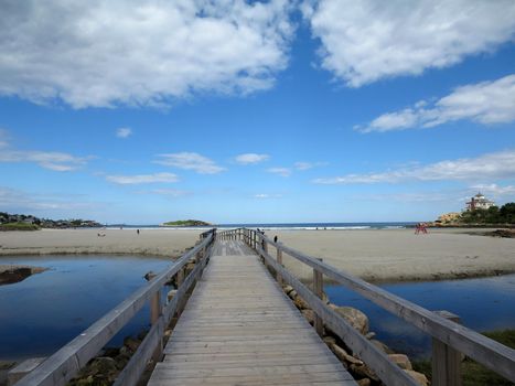 Wood Bridge over Stream leading to beach with people playing in the surf at Good Harbor Beach, Gloucester, Massachusetts. With Lifeguard stand, houses, and Salt Island in the distance.