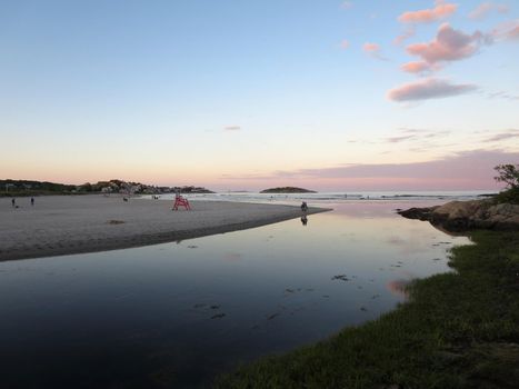 Stream leading to beach with people playing in the surf at Good Harbor Beach, Gloucester, Massachusetts during dusk. With Lifeguard stand and Salt Island in the distance, beautiful pink light.