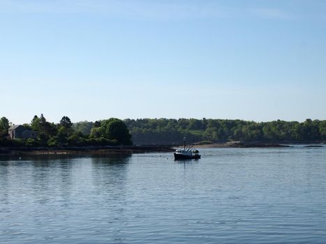 Fishing boat rests in water of Casco Bay off shore of island with house and trees in Maine.