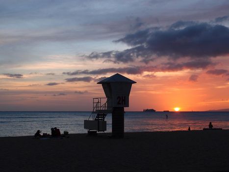 Dramatic sky lighting of sunset on San Souci Beach with stand-up paddle boarder, boats in the ocean, lifeguard stand, and people watching sunset in Waikiki on Oahu, Hawaii. 